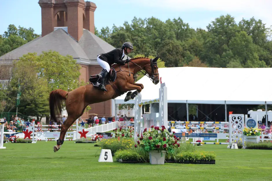 A brown horse with a rider leaping over a white fence in a vast green field.